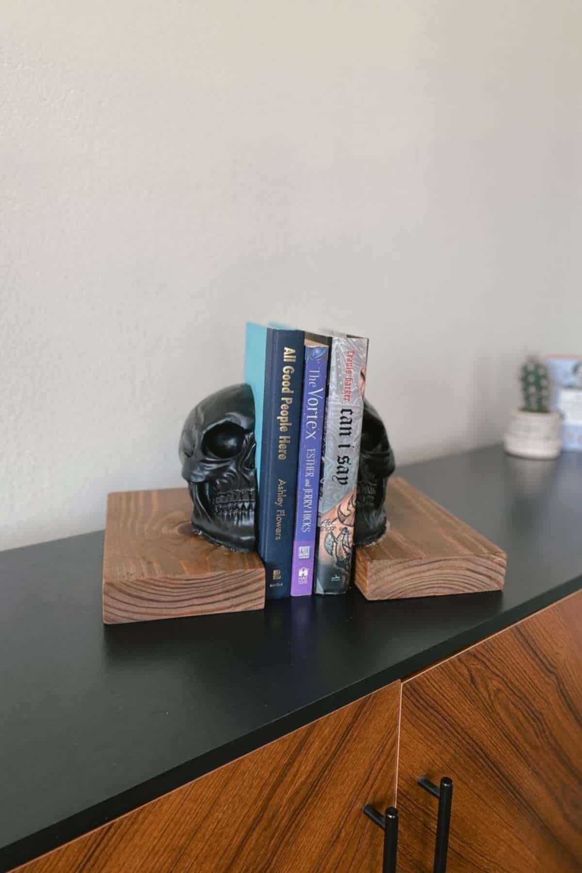Three books are held upright by black skull-shaped bookends on a wooden surface; a small cactus and container are in the background.
