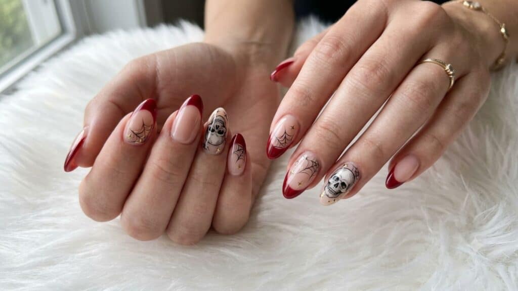 Hands with almond-shaped nails featuring red tips, skull and spiderweb nail art, resting on a white furry surface.