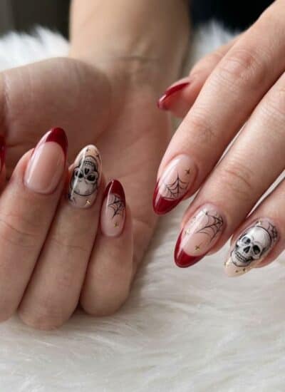 Hands with almond-shaped nails featuring red tips, skull and spiderweb nail art, resting on a white furry surface.
