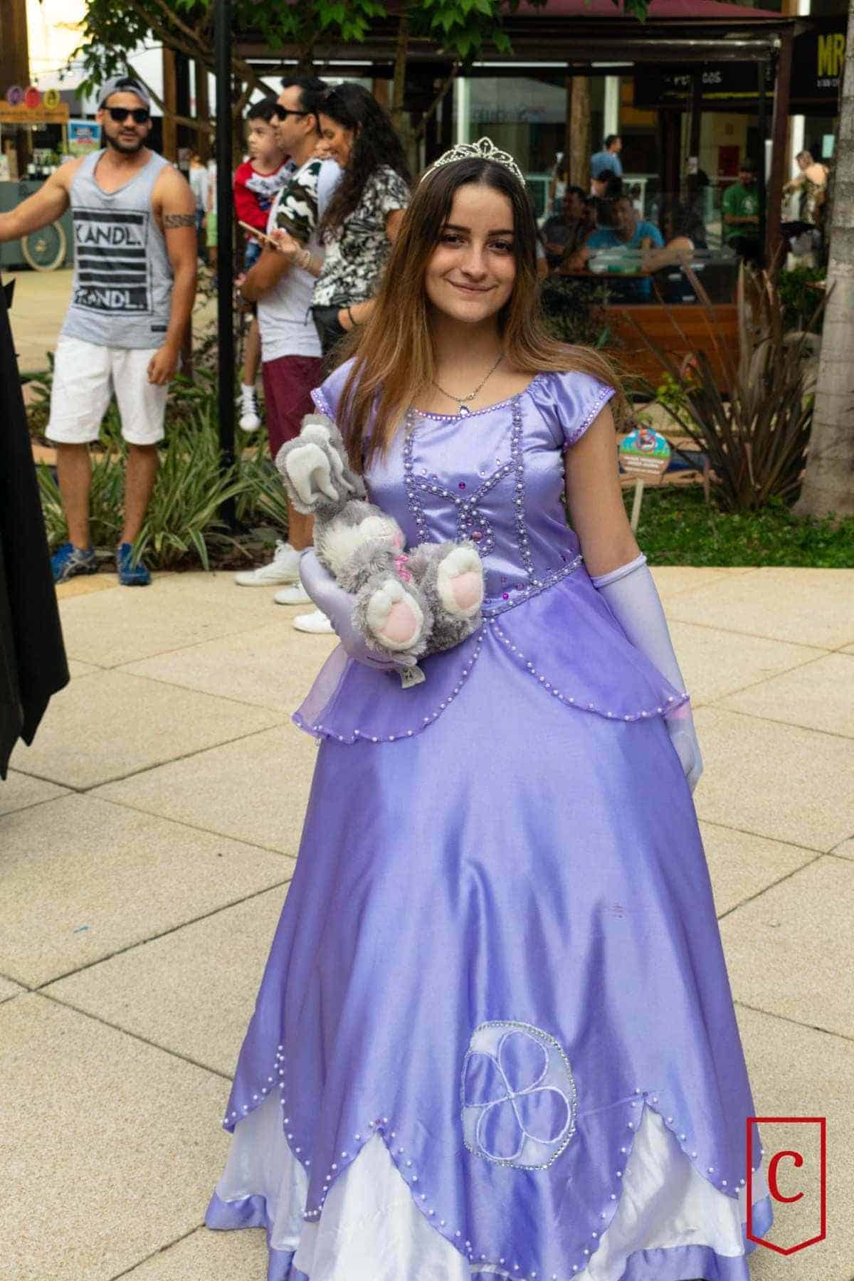 A young woman in a lavender princess dress and tiara holds a stuffed rabbit, standing outdoors among people.