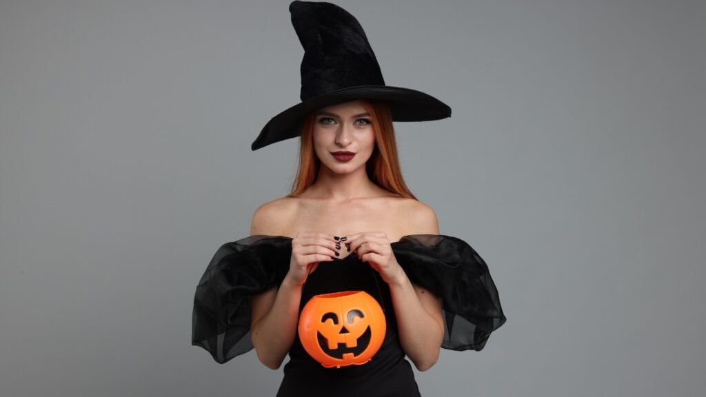 Woman in a black witch costume holds an orange plastic pumpkin bucket, standing against a plain gray background.