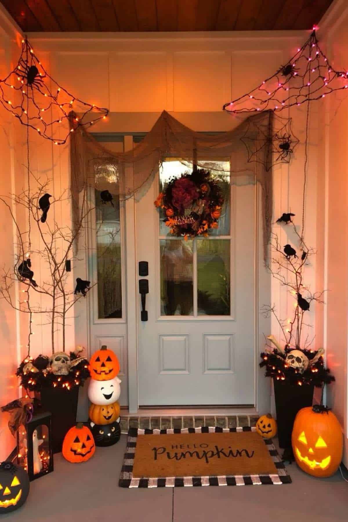 Front porch decorated for Halloween with pumpkins, jack-o'-lanterns, faux spiders, webs, skulls, bats, branches, and a doormat reading "Hello Pumpkin" in front of a closed white door.