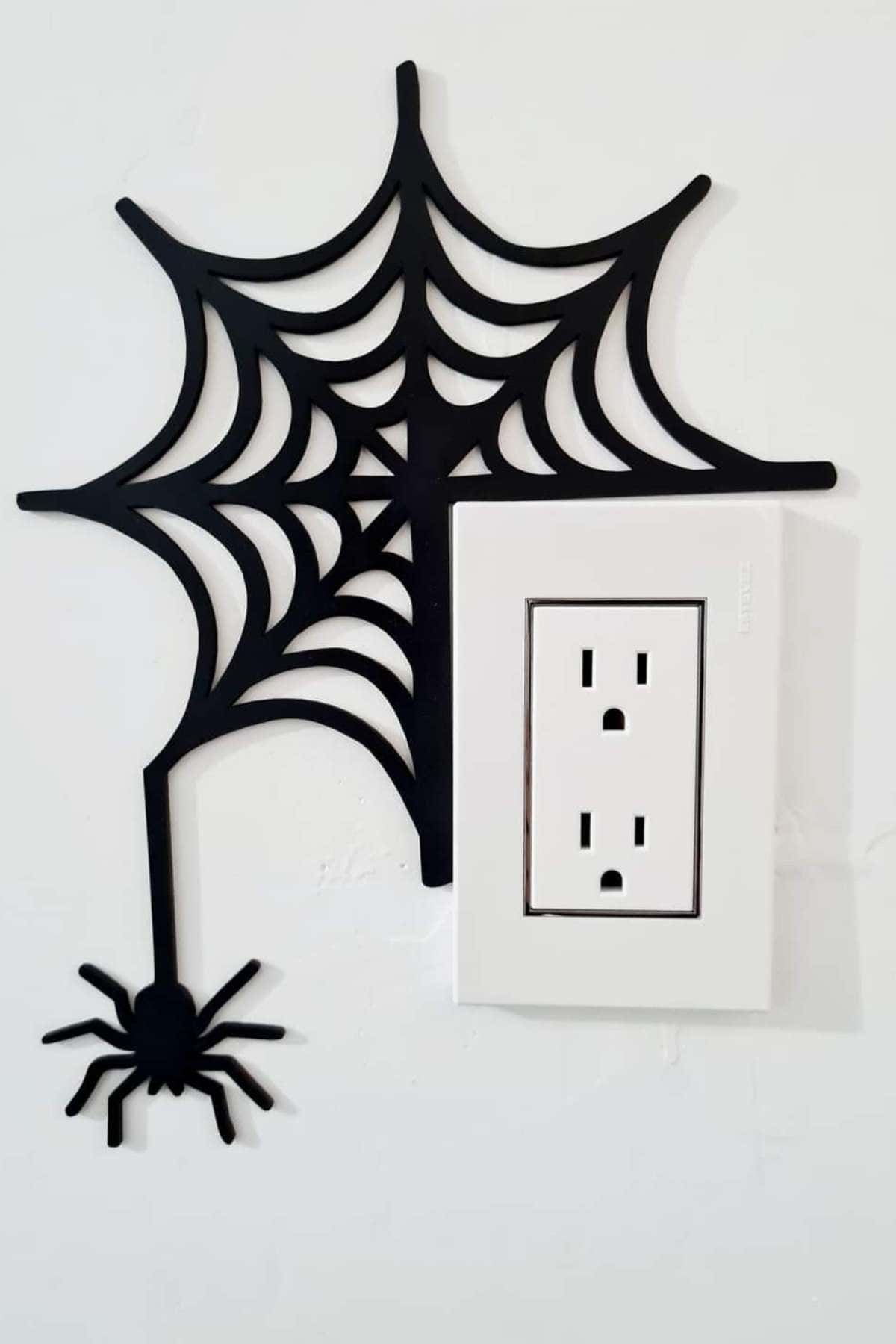 A black spider and web wall decoration is placed next to a white electrical outlet on a plain white wall.