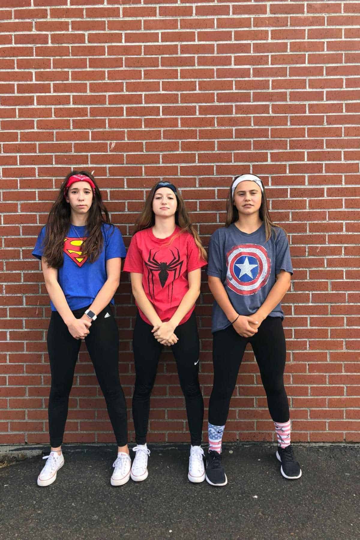 Three young women stand against a brick wall, each wearing a superhero T-shirt: Superman, Spiderman, and Captain America. They are dressed similarly in black pants and sneakers.