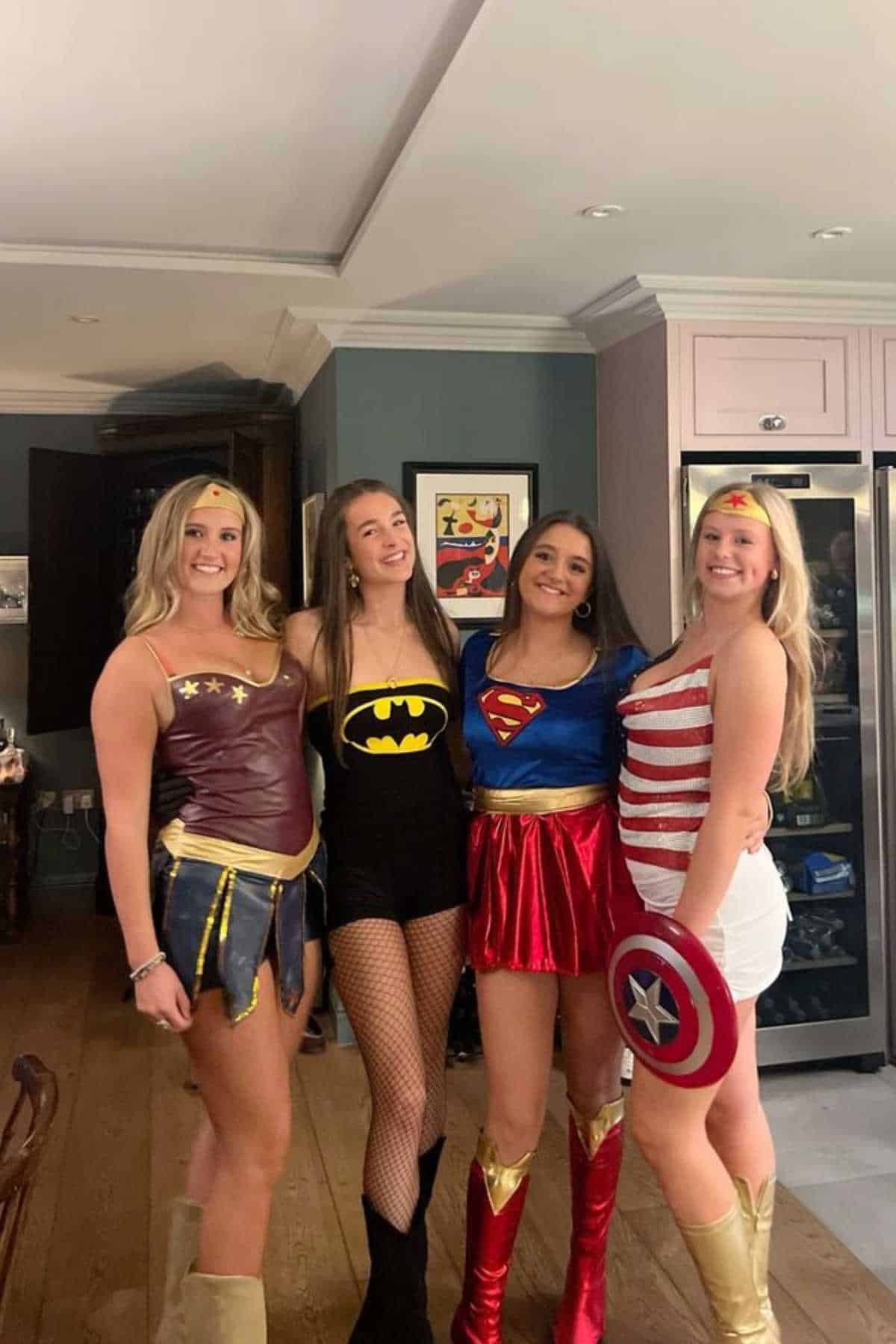 Four women stand indoors, smiling and dressed in superhero costumes representing Wonder Woman, Batman, Supergirl, and Captain America.