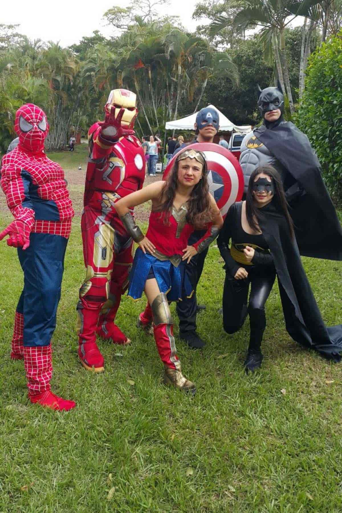 Six people in superhero costumes, including Spider-Man, Iron Man, Wonder Woman, Captain America, Batman, and Batgirl, pose together outdoors on grass with trees and a white tent in the background.