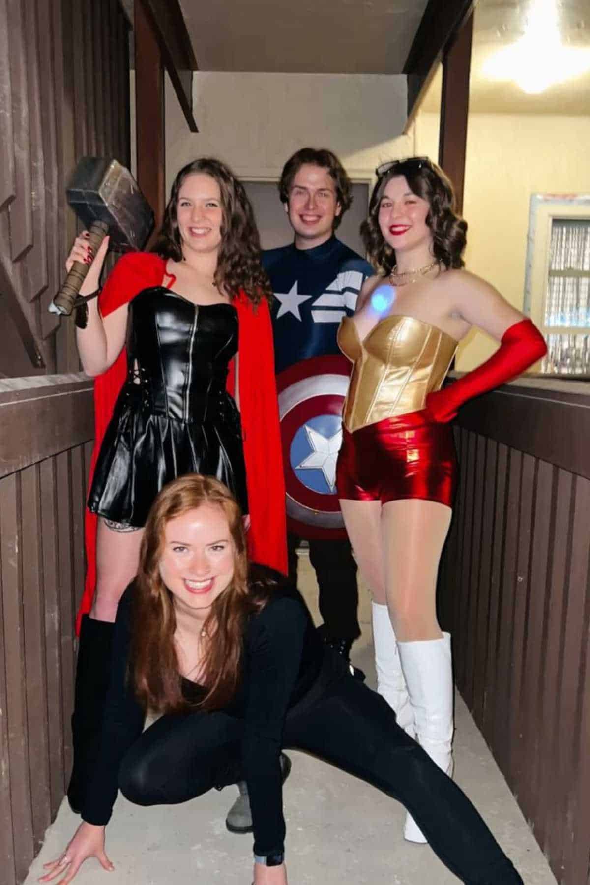 Four people in superhero costumes pose in a hallway; one holds a hammer, one has a shield, one wears a gold and red outfit, and one crouches in black attire.