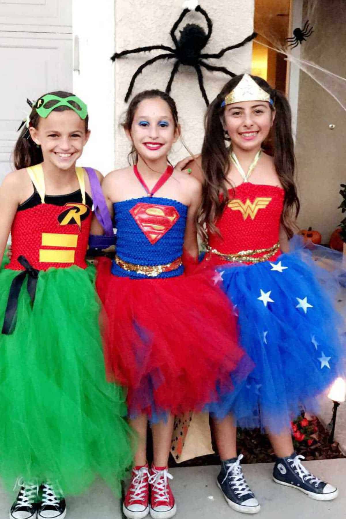 Three girls dressed as Robin, Supergirl, and Wonder Woman stand outside smiling. They wear colorful tutus and sneakers. Halloween decorations with spiders are in the background.