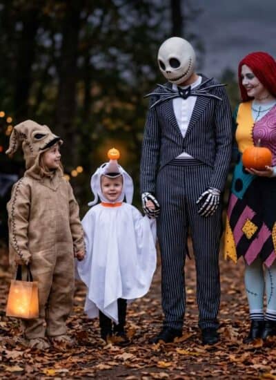 Four people in Halloween costumes stand outdoors surrounded by carved pumpkins and string lights at dusk in a wooded area.