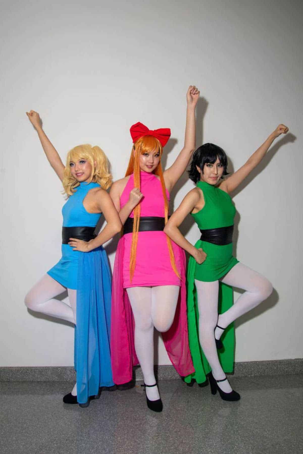 Three people pose in blue, pink, and green costumes with matching wigs, resembling characters from The Powerpuff Girls, standing against a plain light-colored wall.