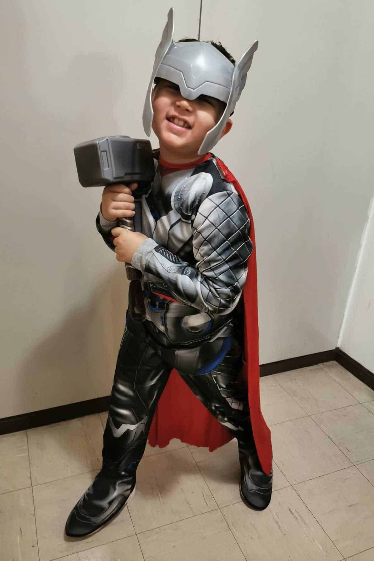 Child in a Thor costume with a helmet, red cape, and holding a toy hammer, standing and smiling indoors on a tiled floor.