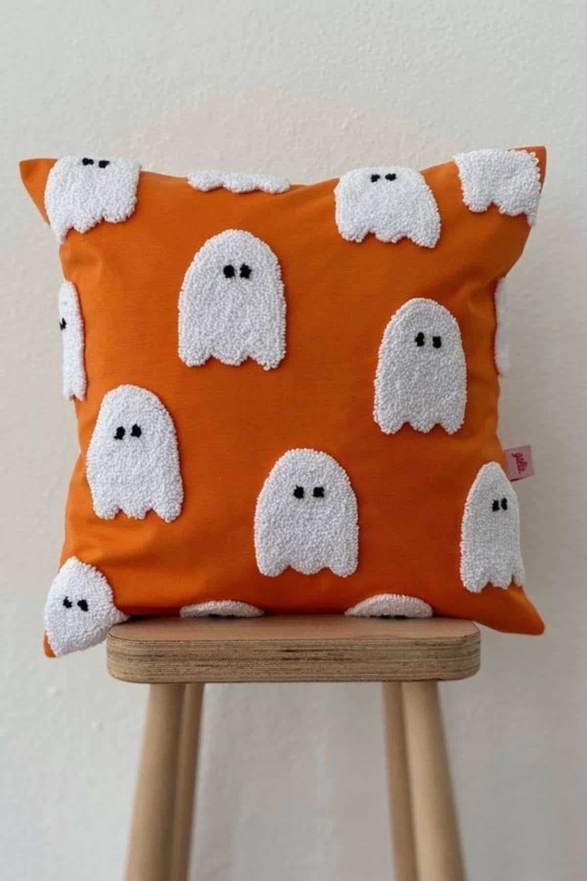 An orange pillow with white ghost patterns featuring black eyes, placed on a wooden stool against a light-colored wall.