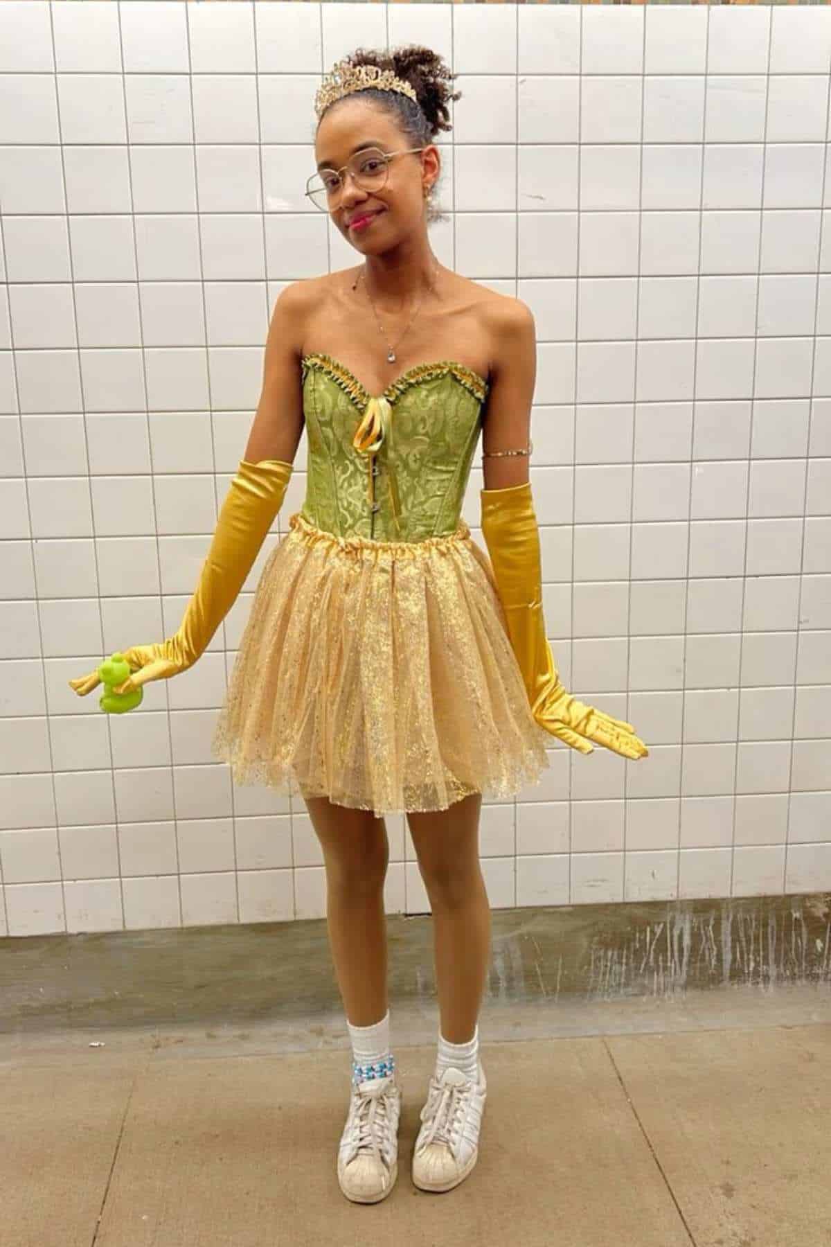Person in a green corset, gold tutu, yellow gloves, sneakers, and a tiara stands in front of a tiled wall, holding a small plush frog.