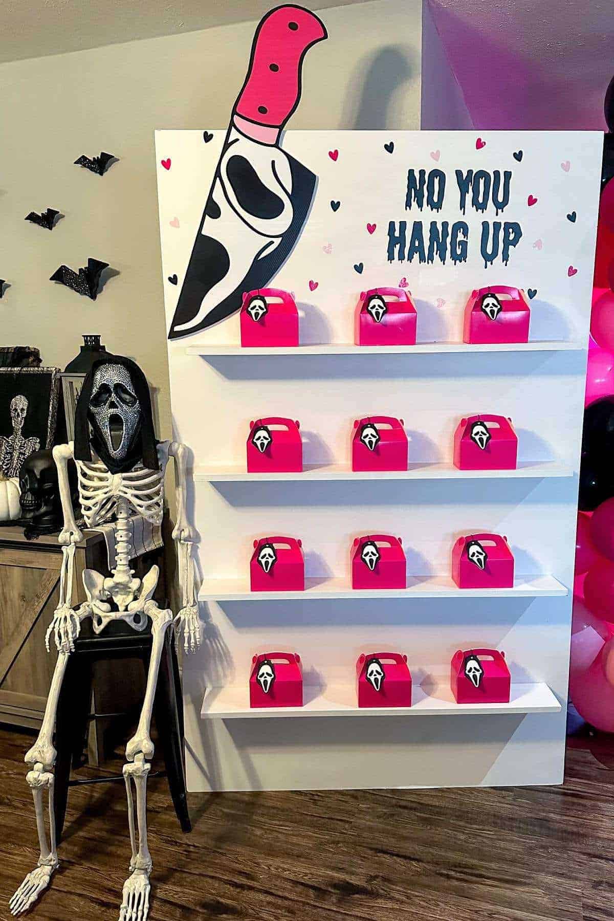 A skeleton in a Ghostface mask sits beside a display with pink boxes, decorated with hearts and the phrase "NO YOU HANG UP" in bold letters.
