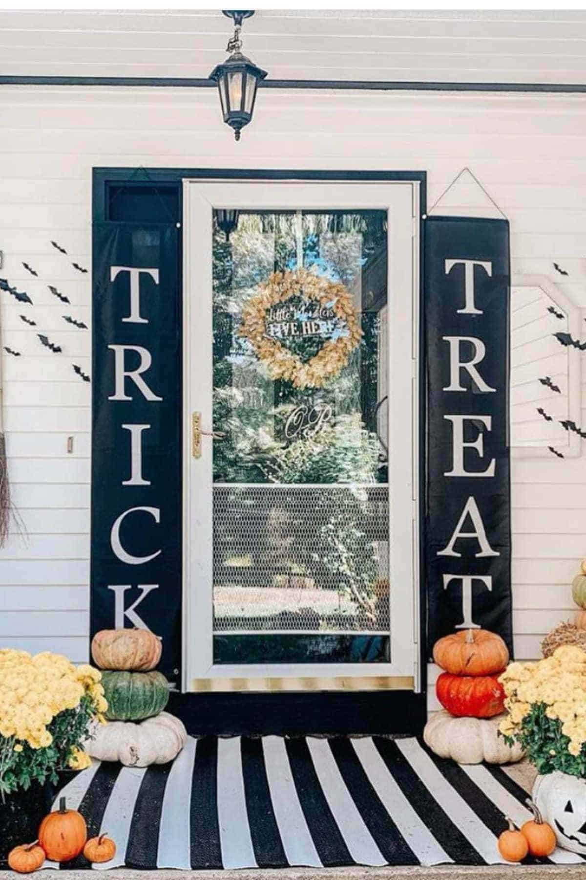 Front porch decorated for Halloween with "TRICK" and "TREAT" signs, pumpkins, yellow flowers, bat cutouts on the wall, and a wreath on the door.