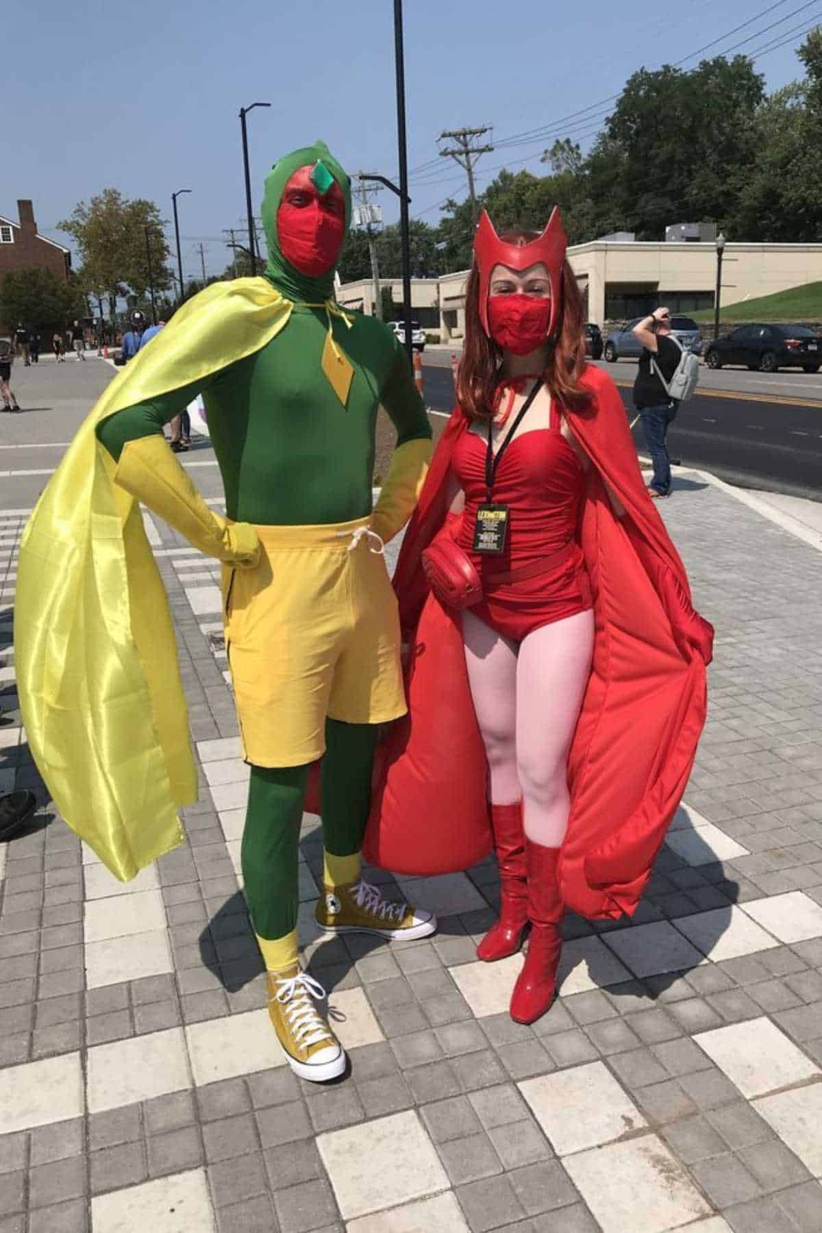 Two people in superhero costumes stand outside on a paved area; one wears green and yellow, the other wears red with a cape and headpiece.