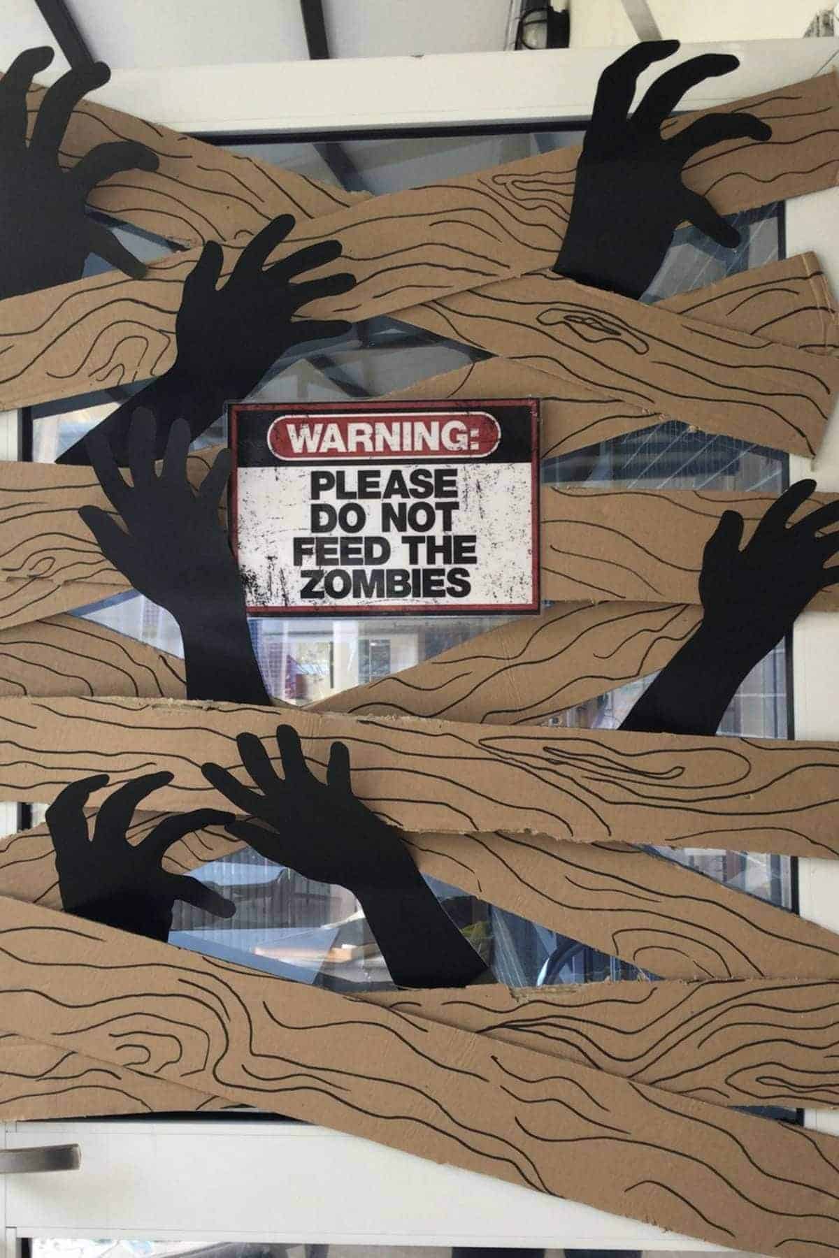 Black cutout hands reach through cardboard strips on a door, with a sign reading "Warning: Please do not feed the zombies" displayed in the center.