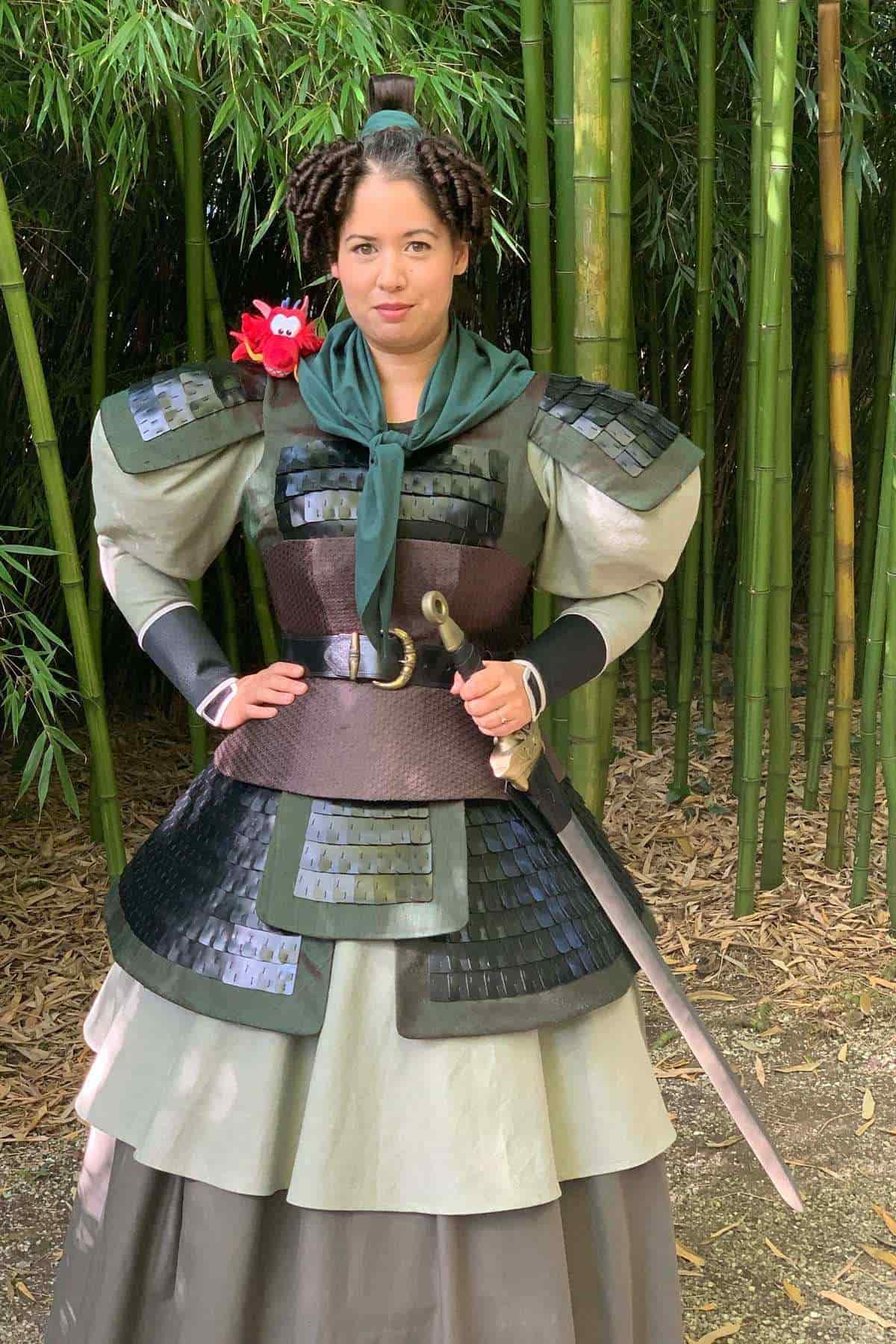 Person in a fantasy warrior costume stands in front of bamboo, holding a sword and wearing armor with a green scarf and a red plush toy on their shoulder.