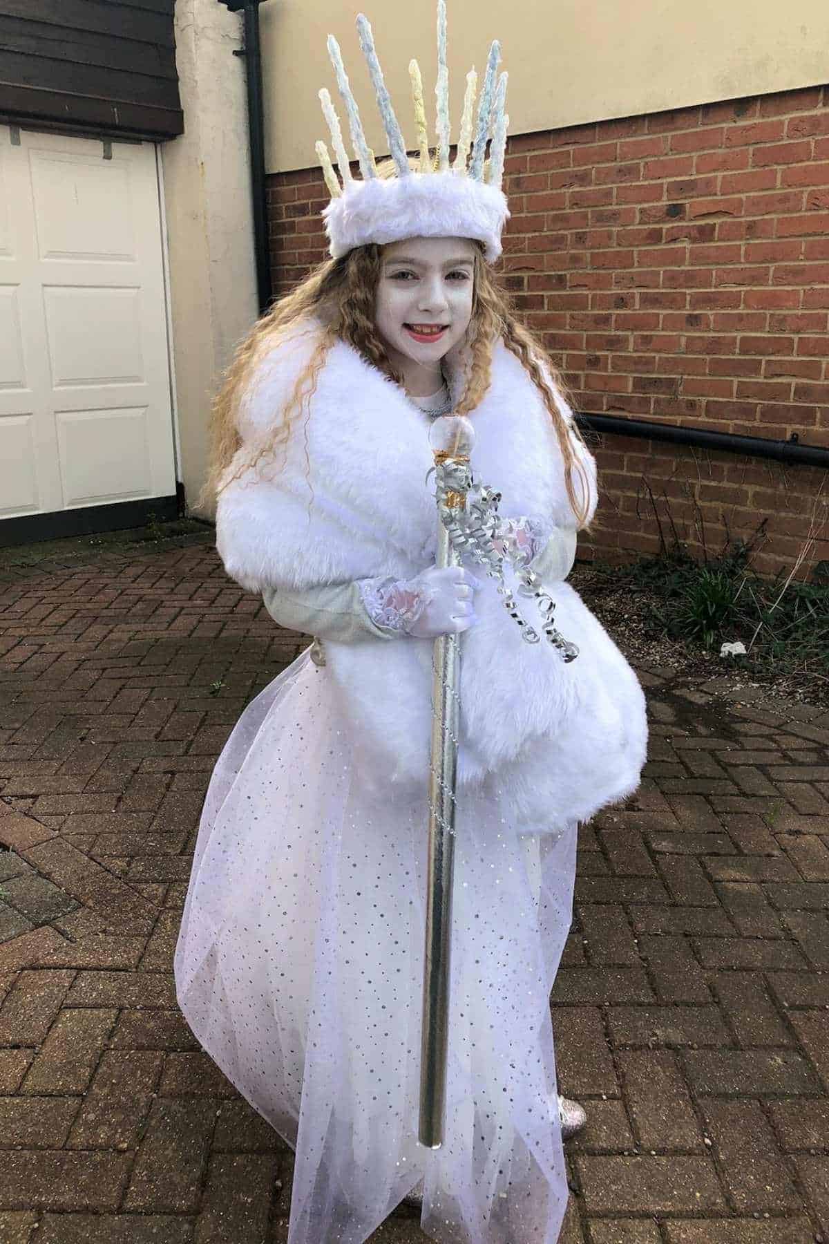Child in a white ice queen costume with a crown and cape, holding a silver scepter, standing outside on a paved area near a brick wall and white garage door.