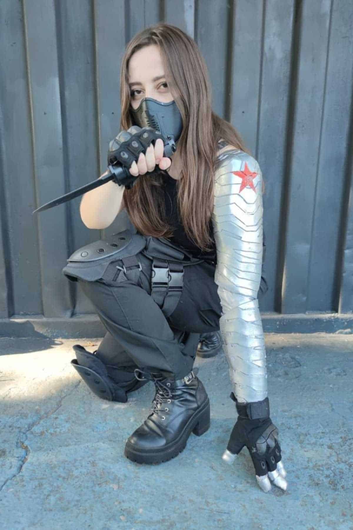Person in black tactical gear and mask crouches, pointing a knife forward. Their left arm is metallic with a red star, resembling a cybernetic limb. Background is a dark metal wall.