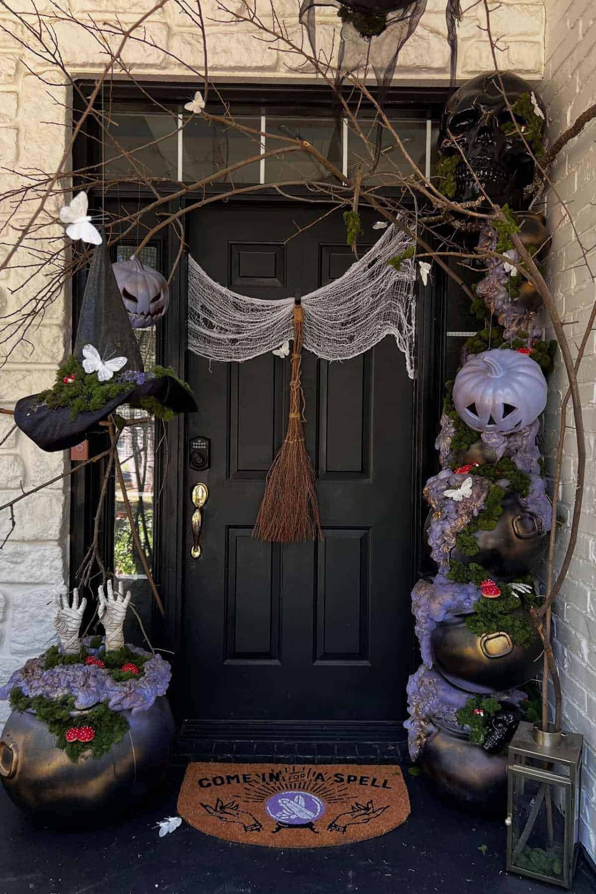 A black door is decorated for Halloween with white pumpkins, cauldrons, a broom, witch hands, a hat, spiderwebs, and a mat that reads “Come in for a spell.”.