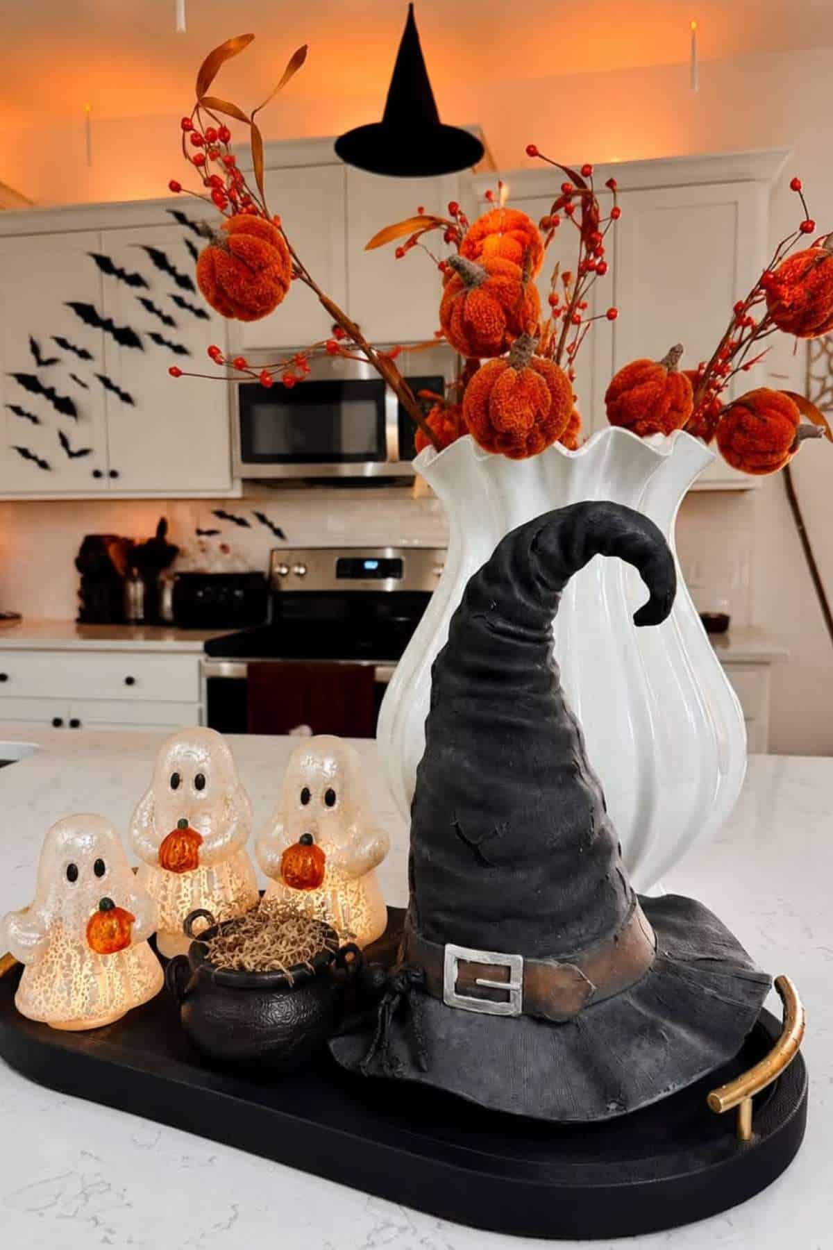 A black witch hat, ceramic cauldron, and three ghost figurines with pumpkins are arranged on a tray. An orange floral arrangement and Halloween bat decals decorate a white kitchen in the background.