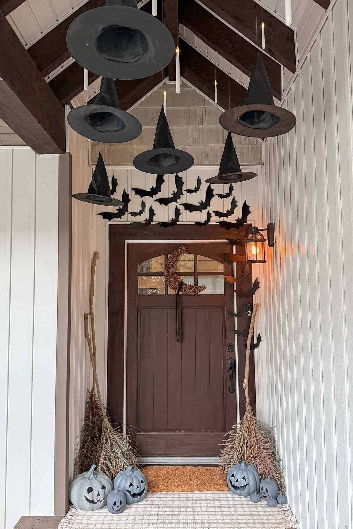 Five black witch hats and paper bats hang above a wooden front door, flanked by twig brooms and painted pumpkins with carved faces, creating a Halloween-themed entryway.