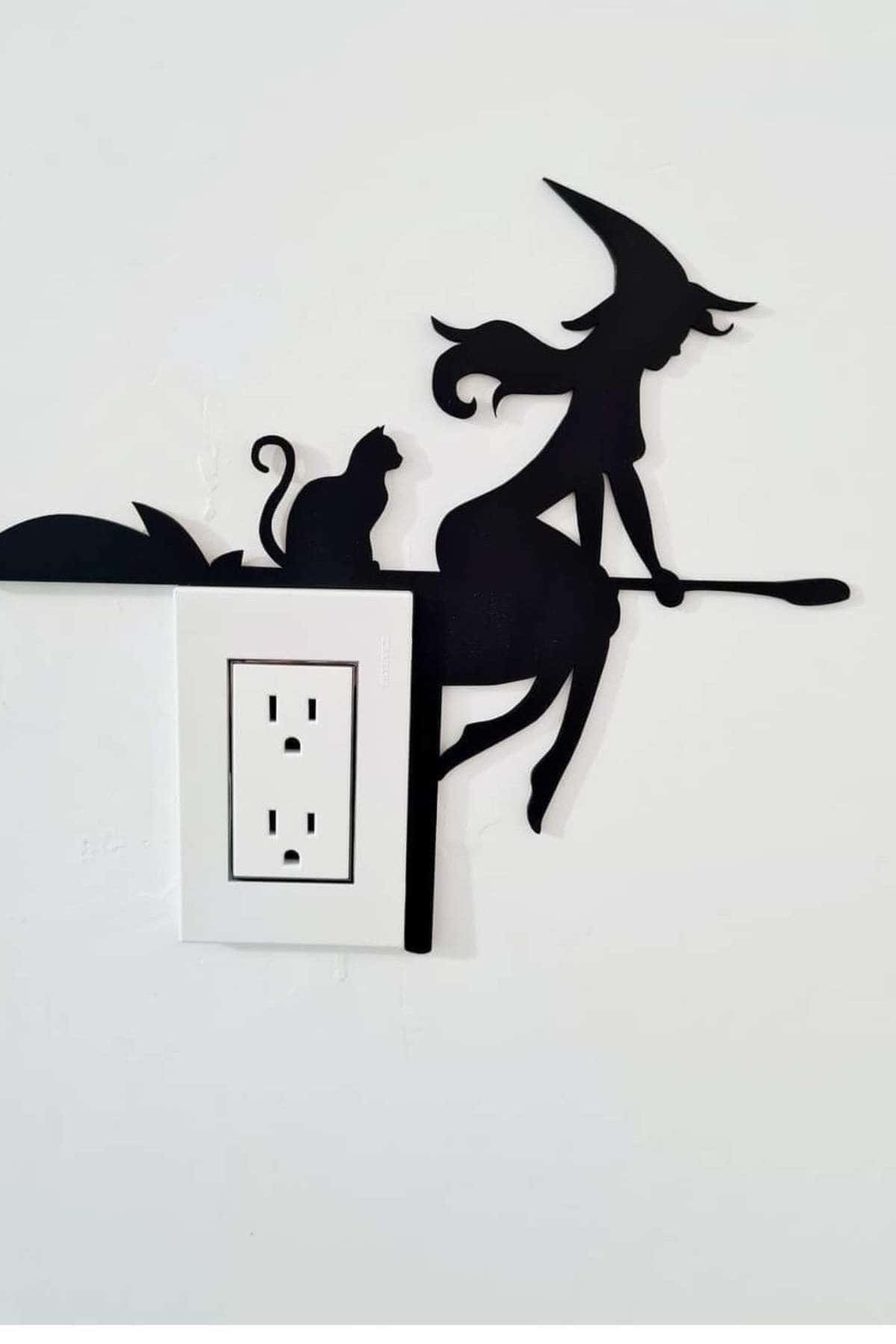Black silhouette wall decal of a witch on a broom with a cat, placed above a power outlet to create a whimsical scene.