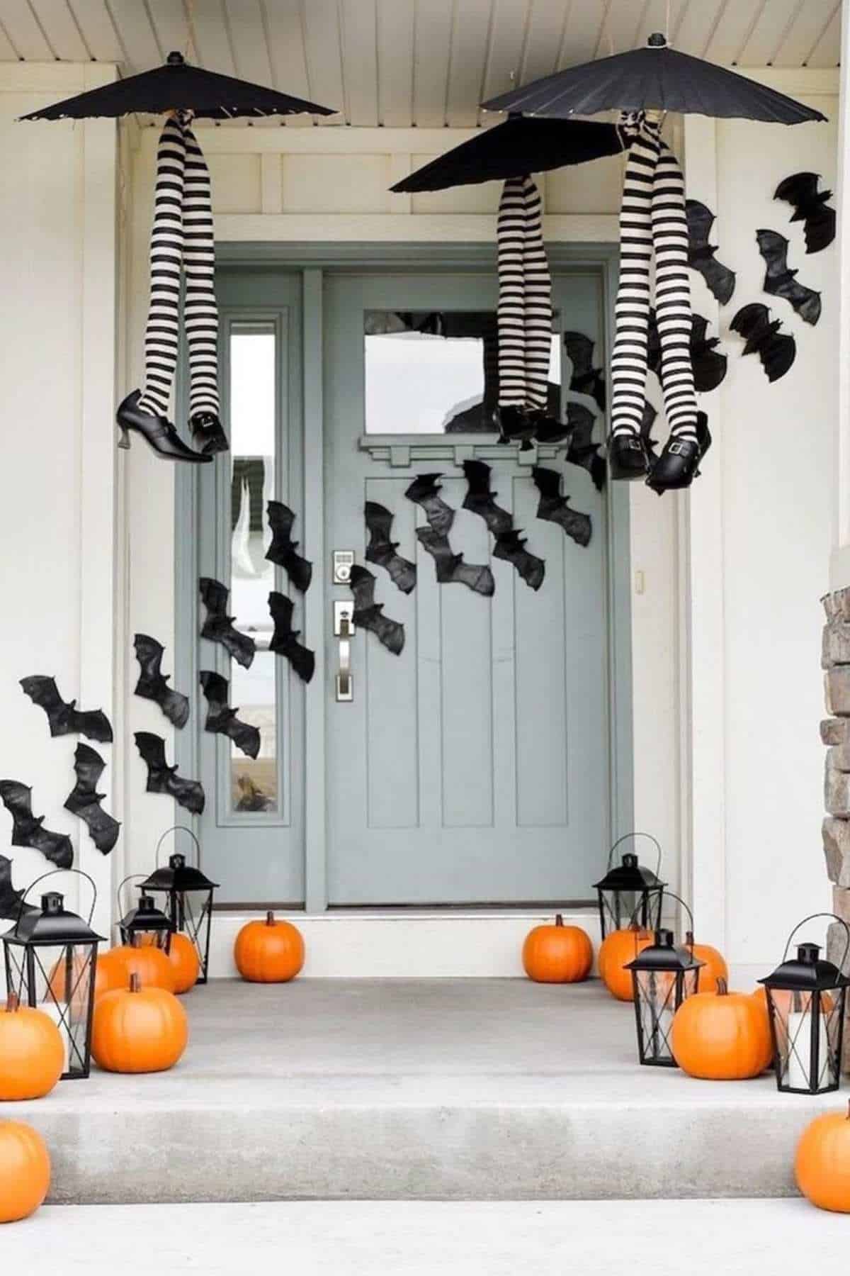 Front porch decorated for Halloween with black lanterns, pumpkins, black paper bats, and two umbrellas holding striped witch legs hanging upside down by the door.