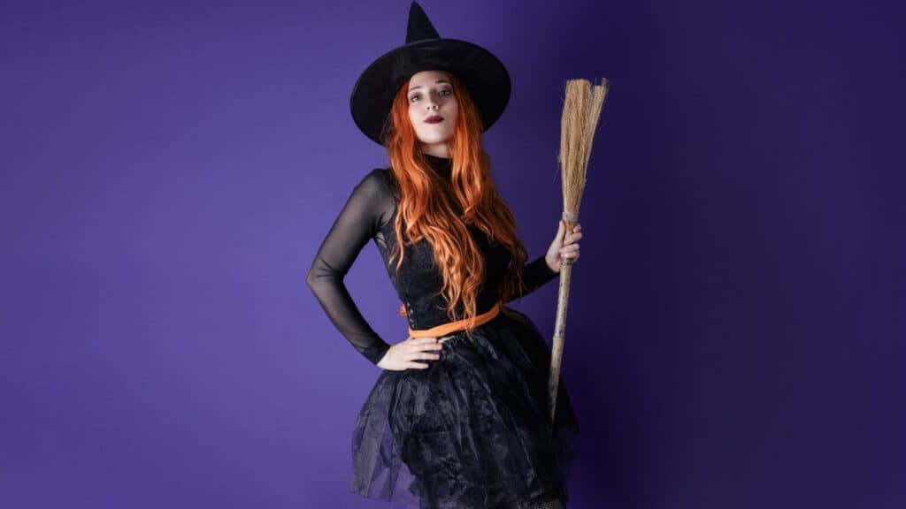 Person in a black witch costume with a pointy hat and long orange hair holds a broom against a solid purple background.