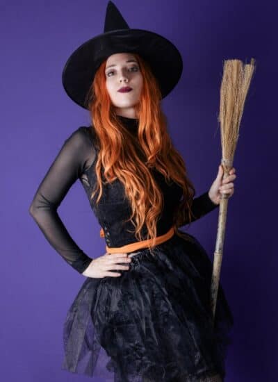 Person in a black witch costume with a pointy hat and long orange hair holds a broom against a solid purple background.