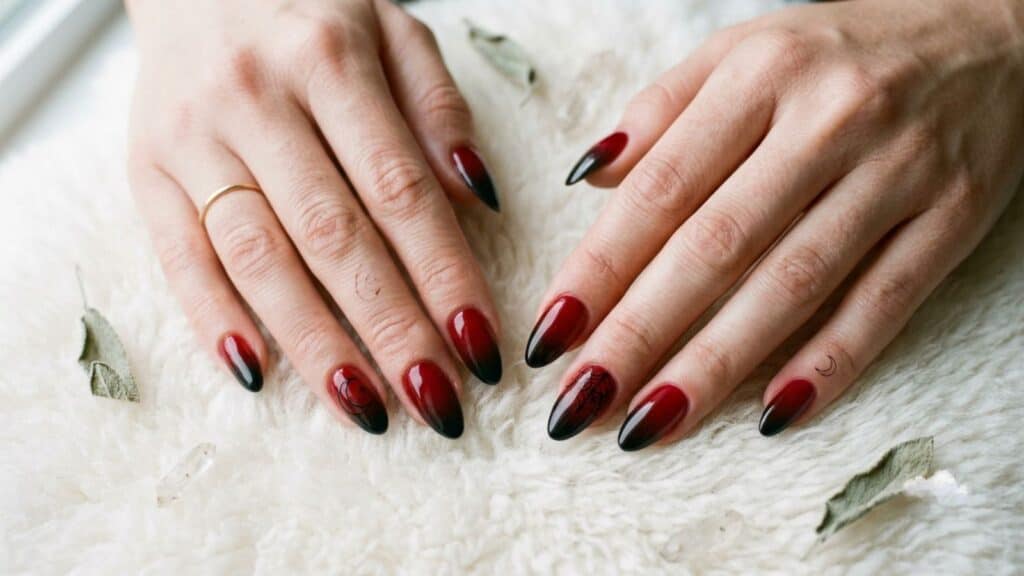 Two hands with long, almond-shaped nails featuring a red and black gradient manicure rest on a white textured surface.