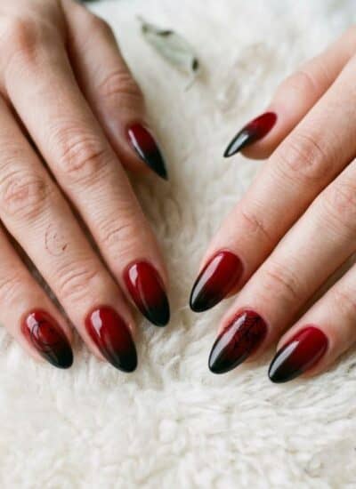 Two hands with long, almond-shaped nails featuring a red and black gradient manicure rest on a white textured surface.