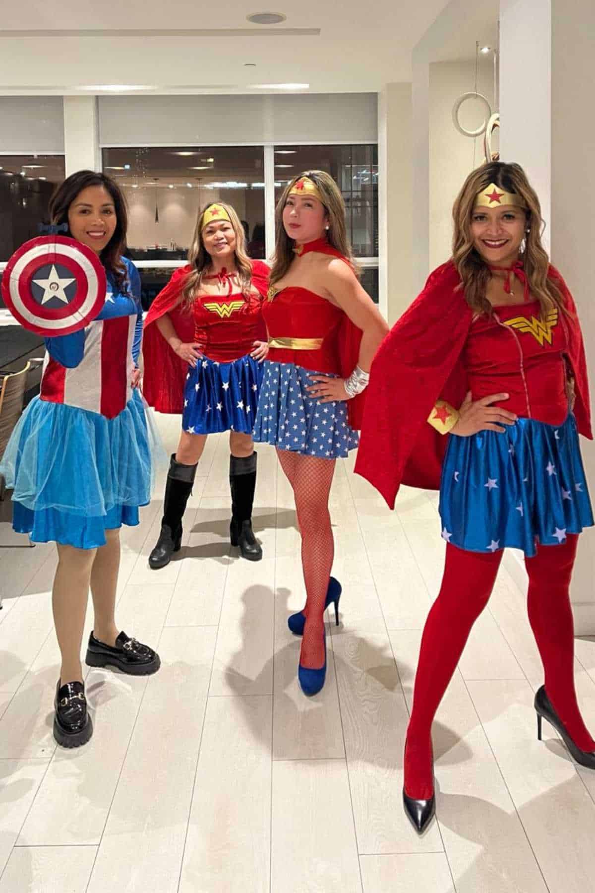 Four women in superhero costumes, including Captain America and Wonder Woman, stand posing together in a brightly lit indoor space with white floors and walls.