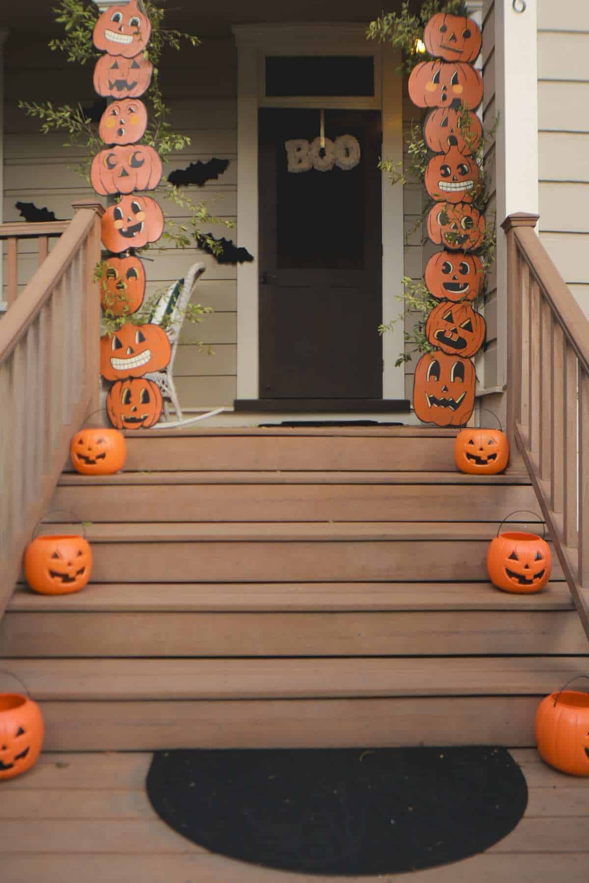 Wooden porch decorated for Halloween with stacked paper jack-o’-lanterns on railings, pumpkin buckets on steps, black bats on walls, and “BOO” sign on the door.