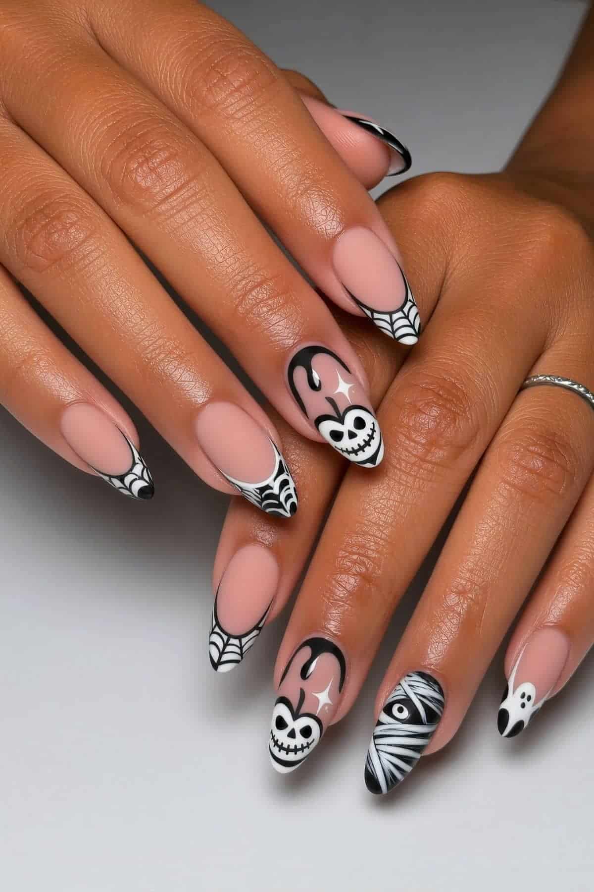 Hands with almond-shaped nails featuring black and white Halloween-themed nail art, including skulls, webs, an eye, and a ghost on a nude base.