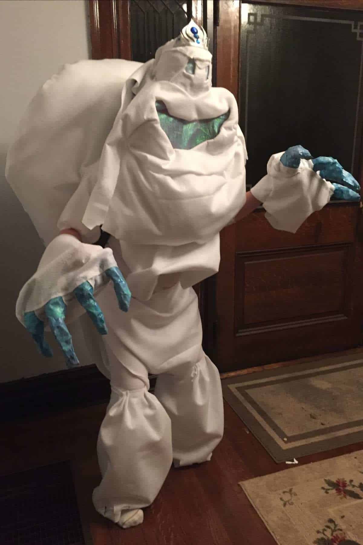 Person wearing a homemade costume resembling a snowy or icy creature with blue hands, standing indoors on a wooden floor near a door.