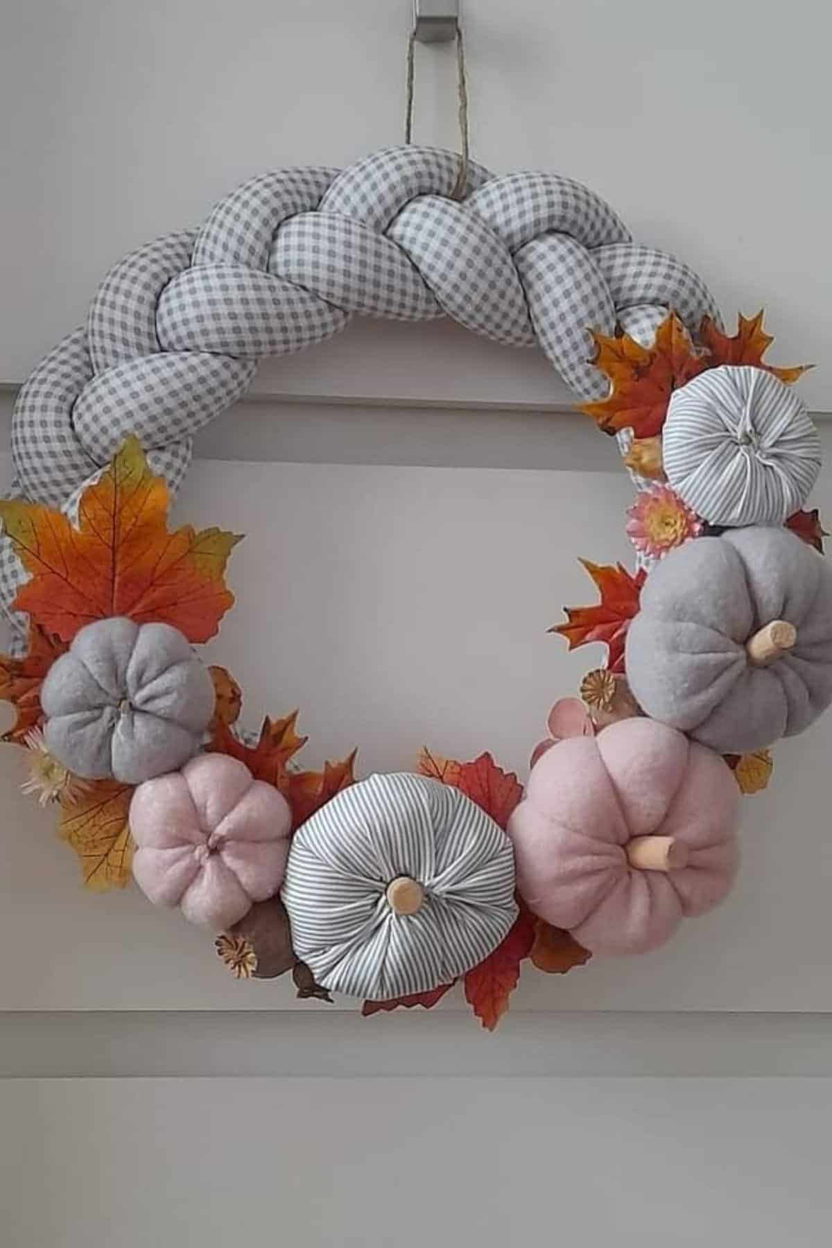 A braided fabric wreath decorated with cloth pumpkins in pink and gray tones, artificial autumn leaves, and small flowers, hanging on a white wall.