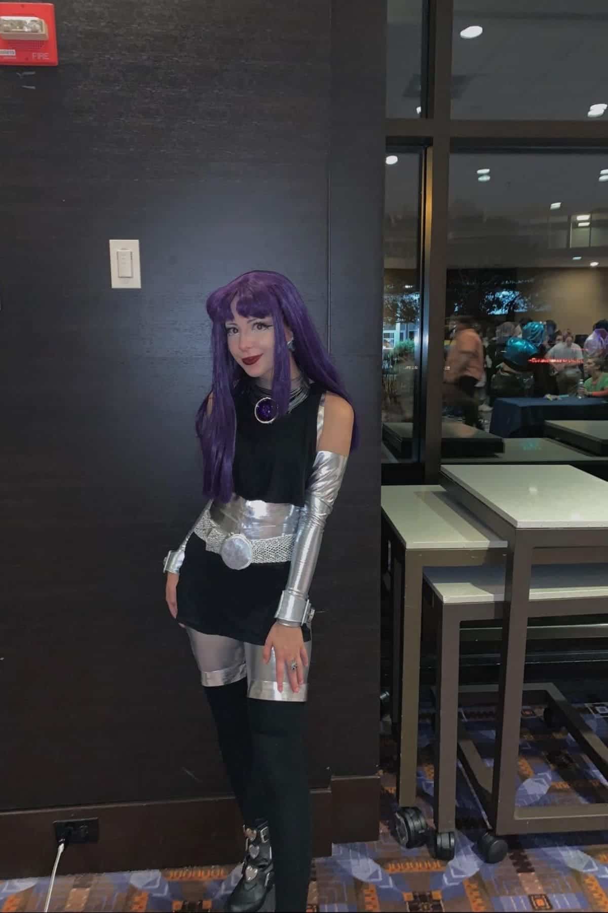 Person dressed in a purple wig and silver costume, posing indoors against a dark wall with tables and people visible in the background.