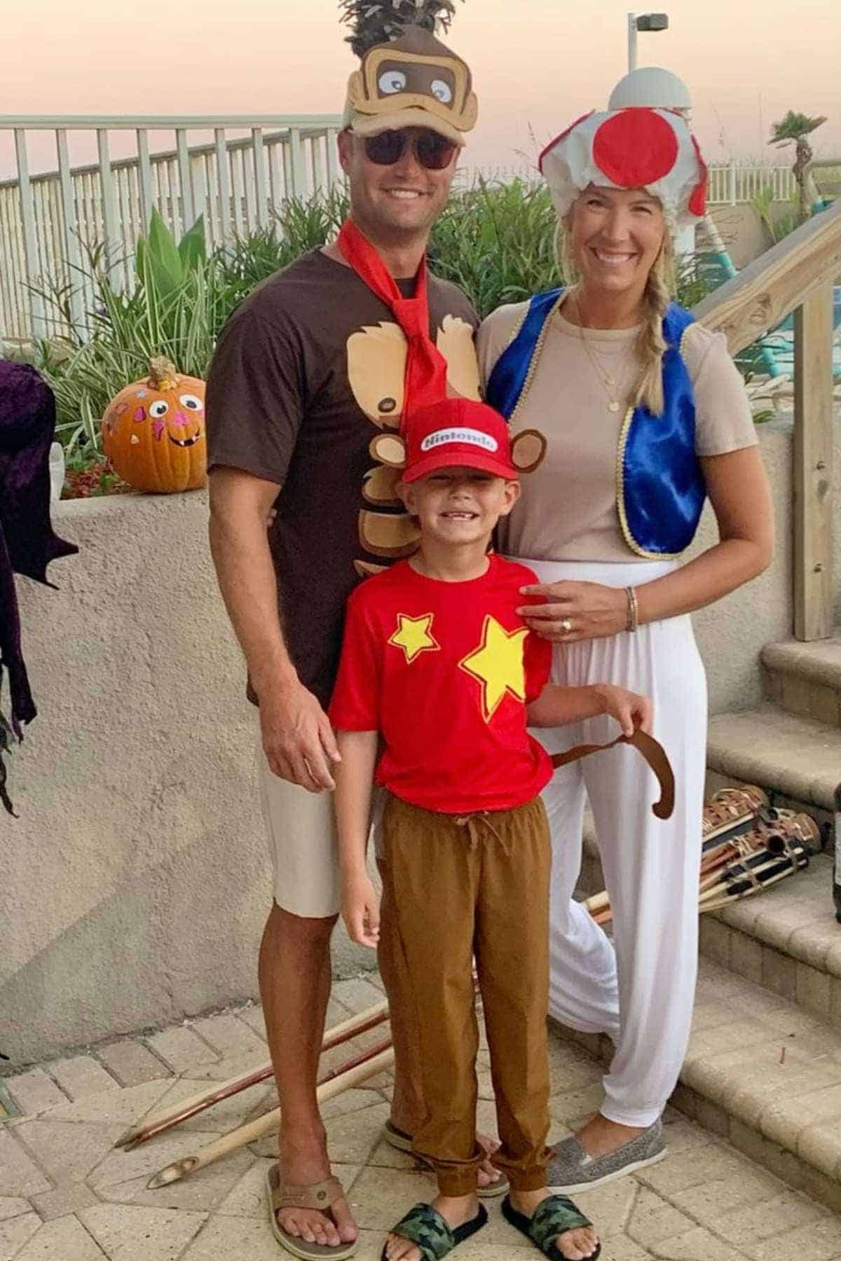 Three people in coordinated costumes pose outside; one dressed as Donkey Kong, one as Diddy Kong, and one as Toad from Mario, with a decorated pumpkin visible on a wall behind them.