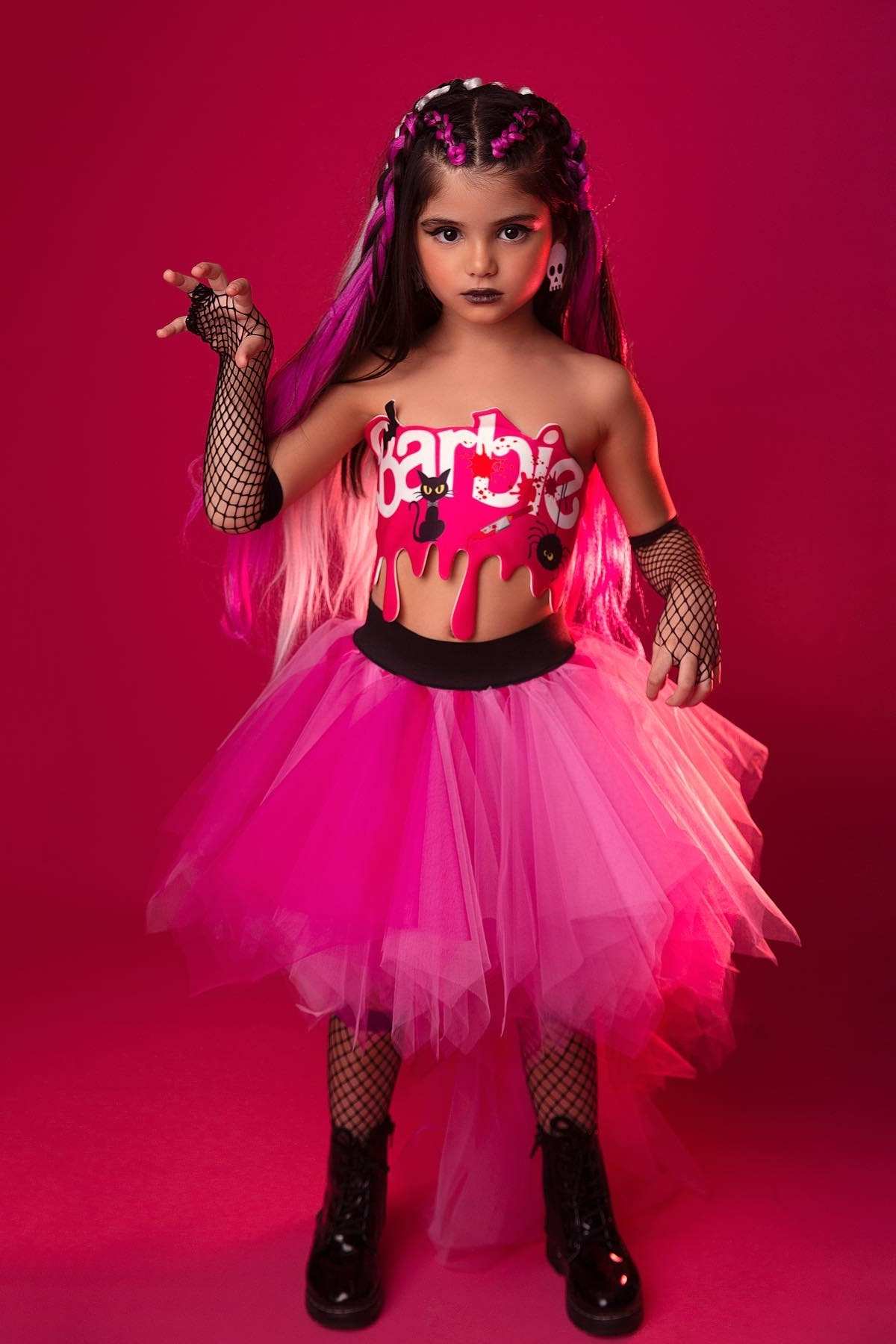 Young girl in a pink and black punk Barbie-themed outfit with a tulle skirt, fishnet gloves, and boots, standing against a solid pink background.