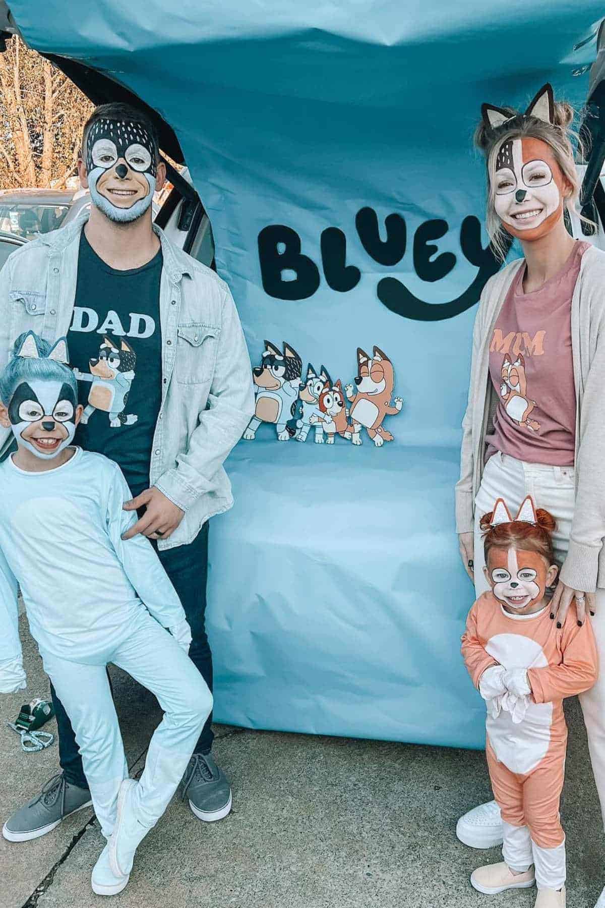 A family of four dressed as Bluey characters poses in front of a Bluey-themed backdrop, with face paint and costumes representing different characters from the show.
