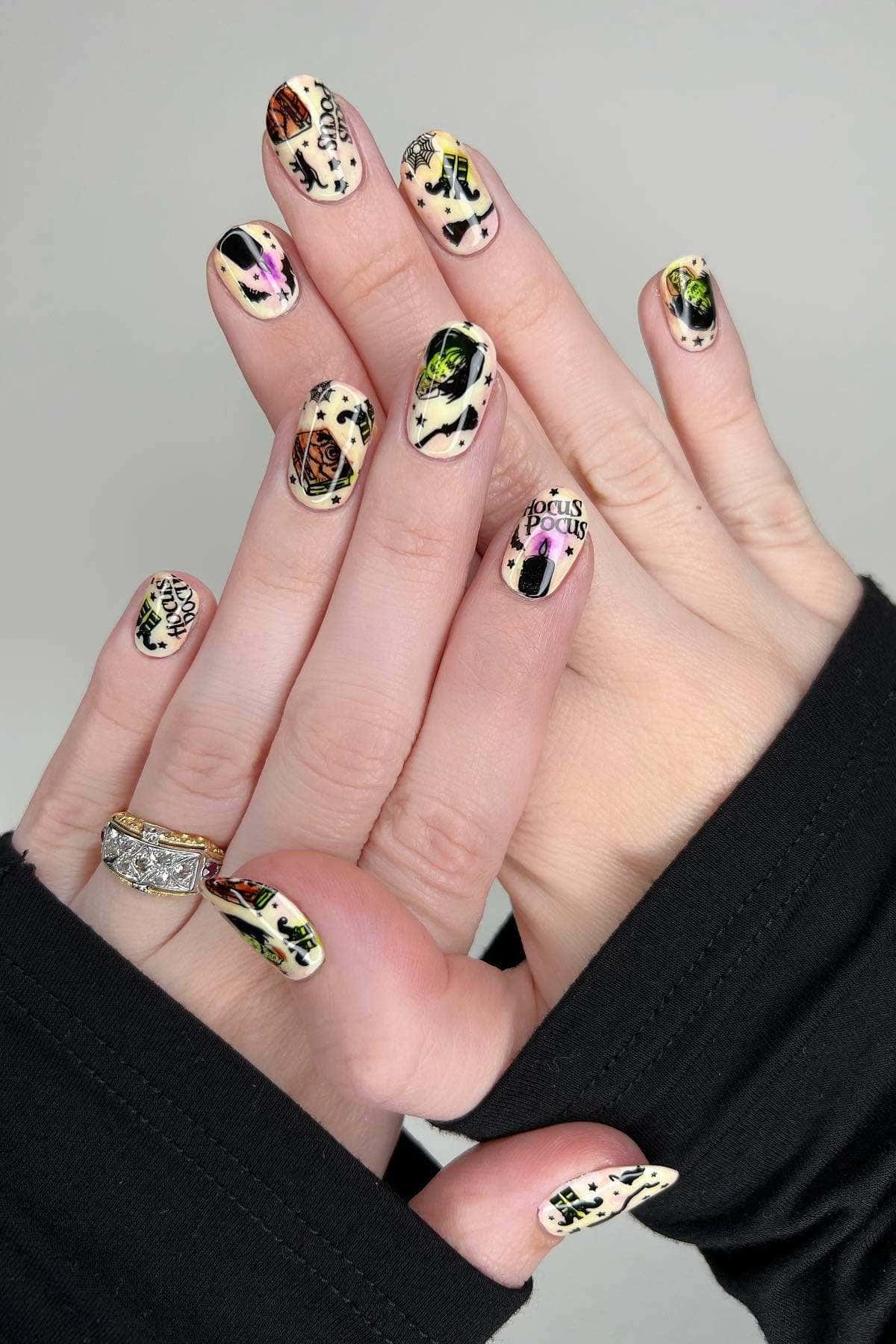 Hands with Halloween-themed nail art featuring black, green, and purple designs, including the words “Hocus Pocus.” One hand has a gold ring. Both hands are partially covered by black sleeves.