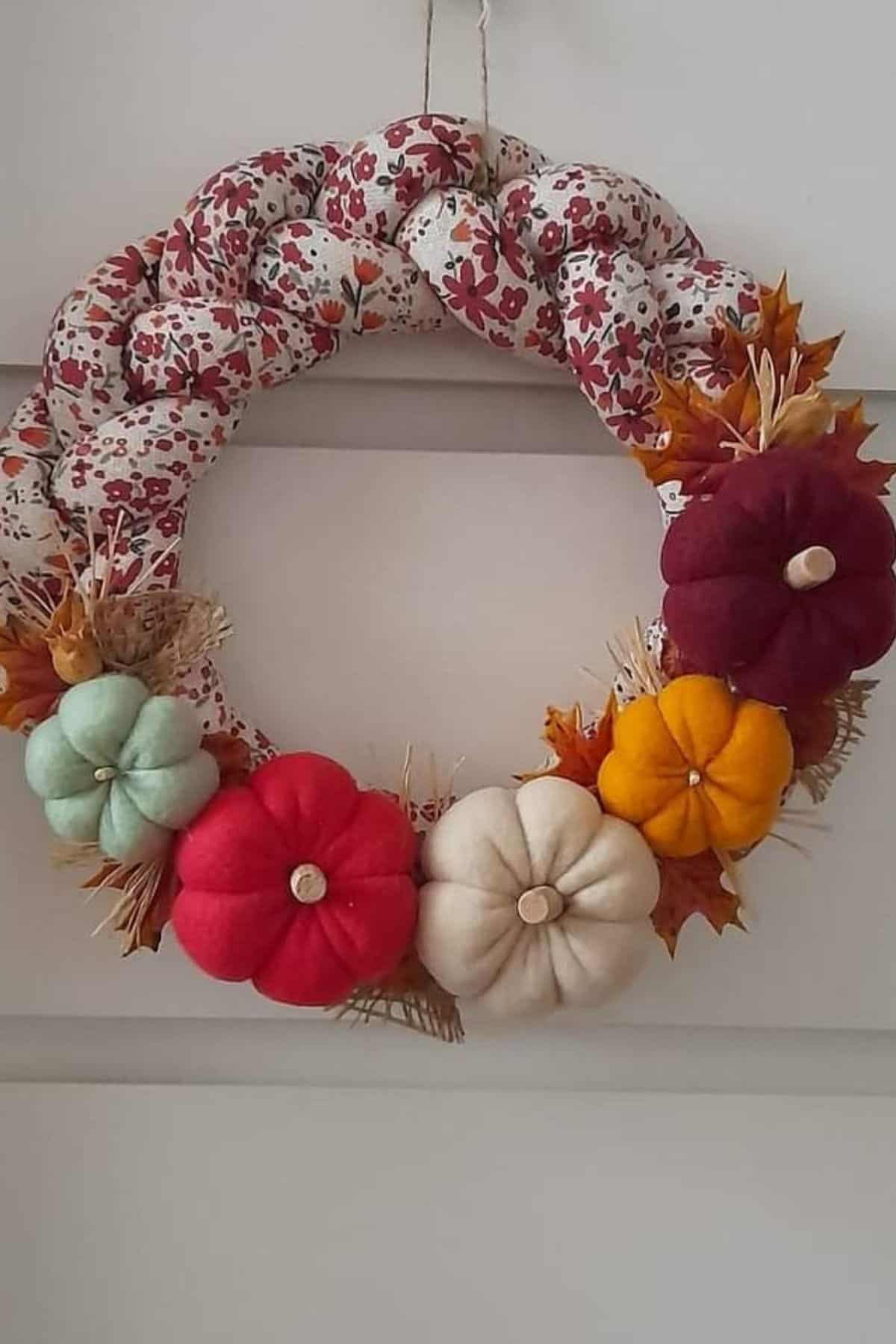 A fabric wreath with a floral pattern is decorated with small, colorful stuffed pumpkins and artificial autumn leaves, hanging on a white wall.