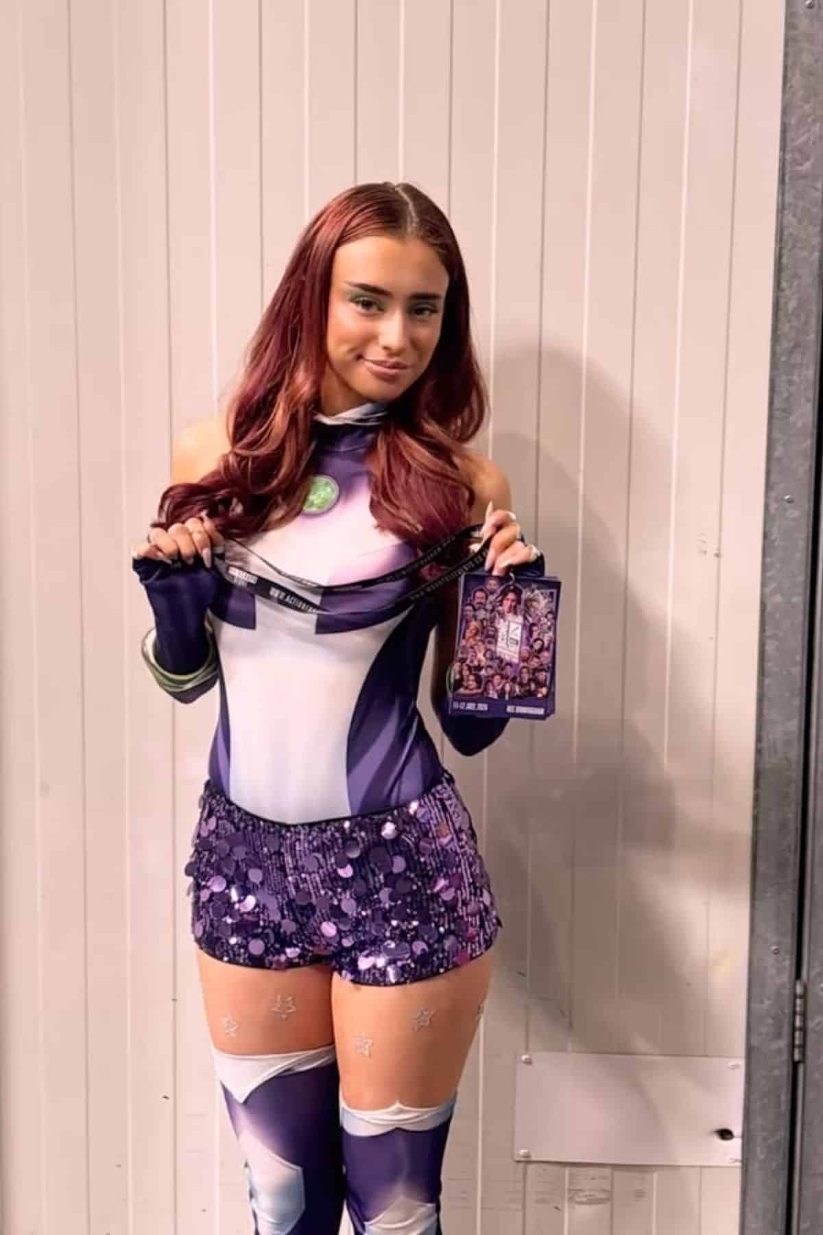 Person in a purple and white superhero costume holding a lanyard and event badge, standing against a vertical white panel wall.