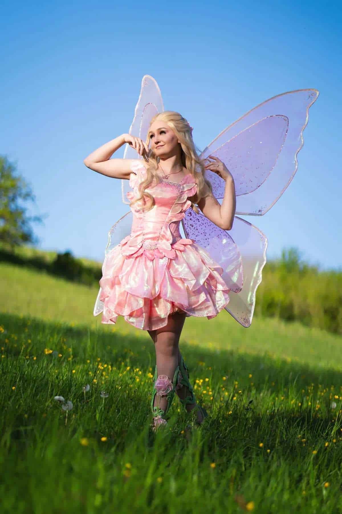 Person in a pink, fairy-inspired costume with large wings stands on green grass under a clear blue sky.