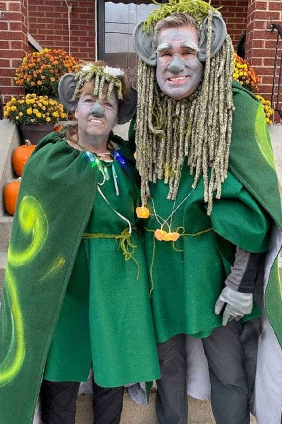 Two people in green costumes with large ears, face paint, and artificial moss stand together on a porch decorated with pumpkins and flowers.
