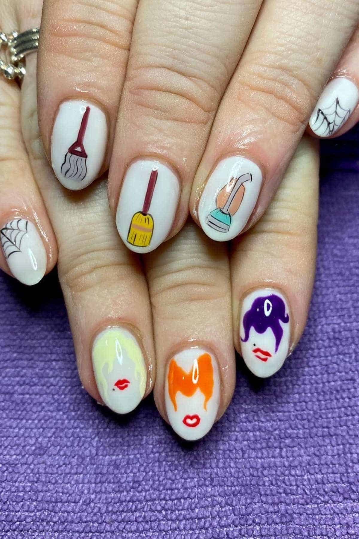 Fingernails painted with Halloween-themed designs, including witches’ faces, a broom, a cauldron, spider webs, and a hat, on a purple textured background.