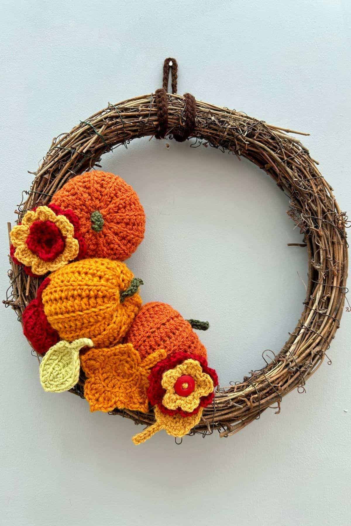 A grapevine wreath decorated with crocheted orange pumpkins and yellow and red flowers, hanging on a plain light-colored wall.