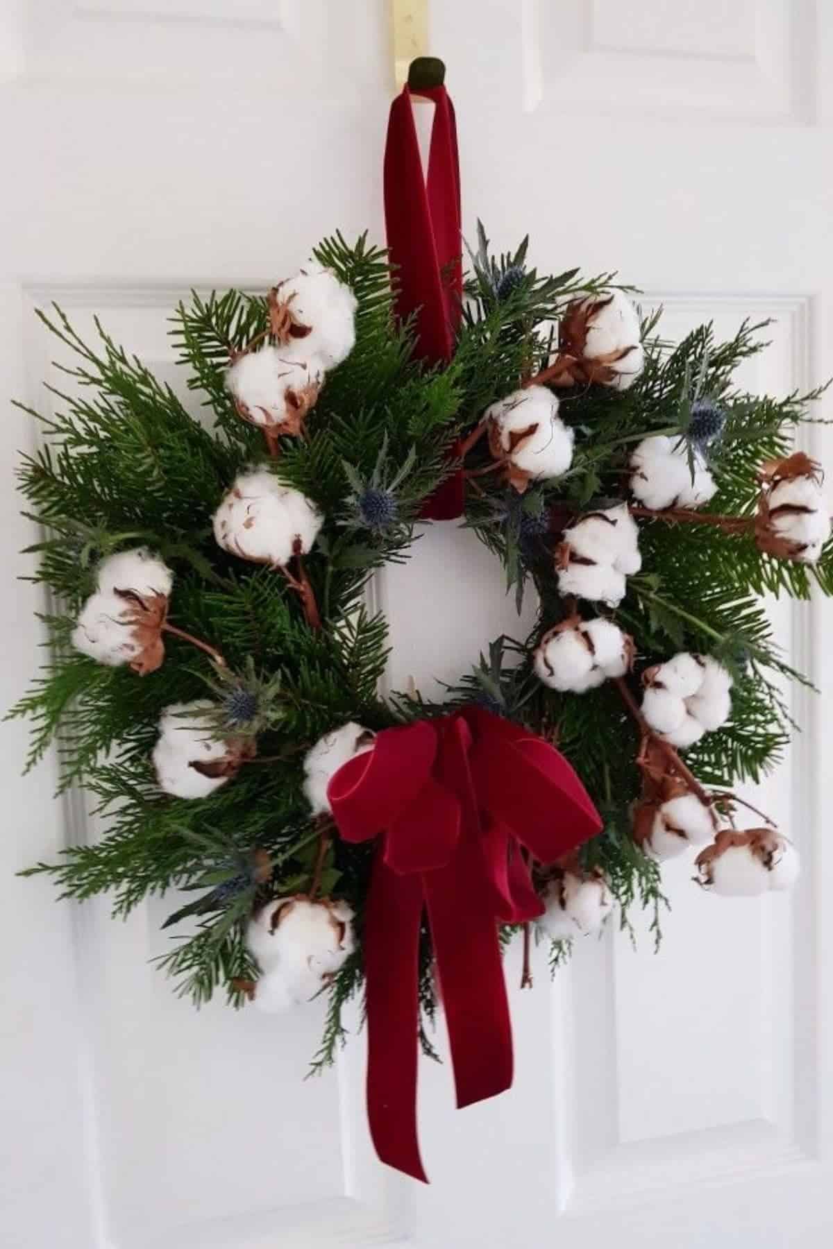 Wreath made of green foliage, cotton bolls, and small thistles, decorated with a red velvet bow, hanging on a white door.